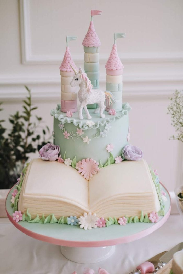 A whimsical cake with a castle and unicorn topper on a pastel blue tier. Below, an open book cake with pastel flowers and green leaves decorates the base. The scene is set on a white and pink rimmed platter.