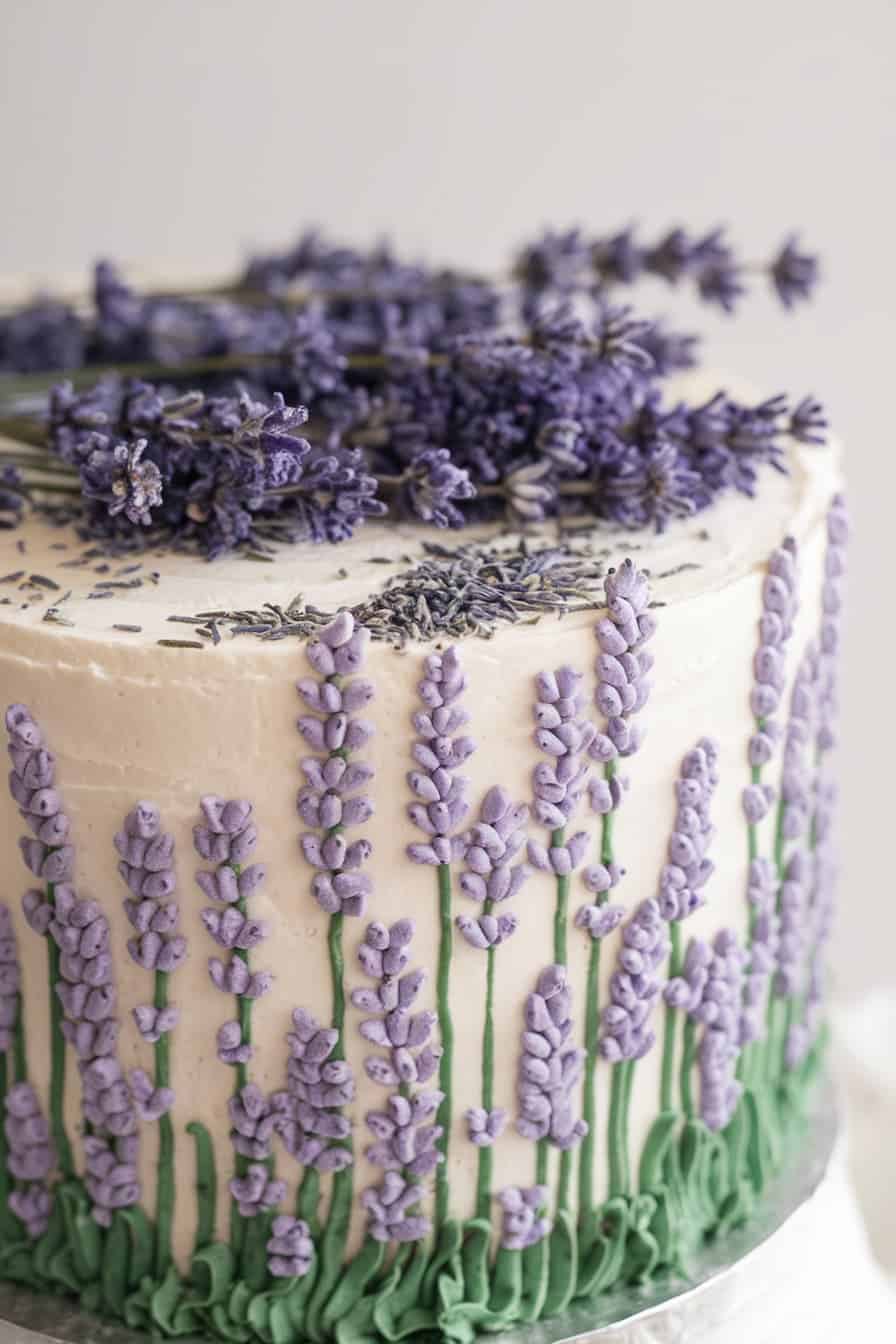 A cake decorated with intricate lavender flowers on a white frosting background. The top is adorned with sprigs of real lavender, adding a natural touch to the elegant design.