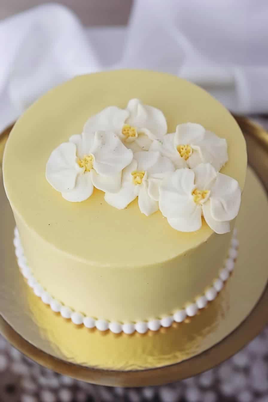 A round yellow cake topped with white edible flowers on a gold platter. The cake has a smooth frosting finish and a border of small white pearls at the base.