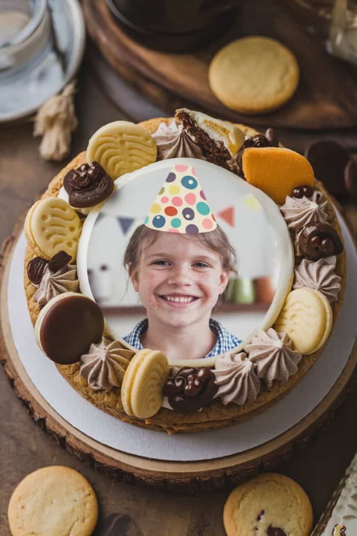 A birthday cake topped with a photo of a smiling child wearing a party hat. The cake is decorated with various cookies, chocolate pieces, and dollops of frosting. A wooden table and teacup are in the background.