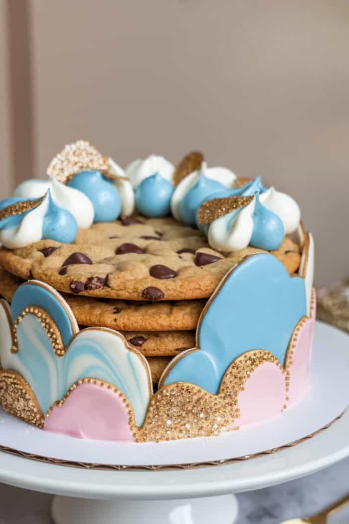 A layered cake made of large chocolate chip cookies, decorated with blue and white whipped cream dollops and gold glitter. The sides feature pastel pink and blue icing with gold accents. The cake sits on a white stand.