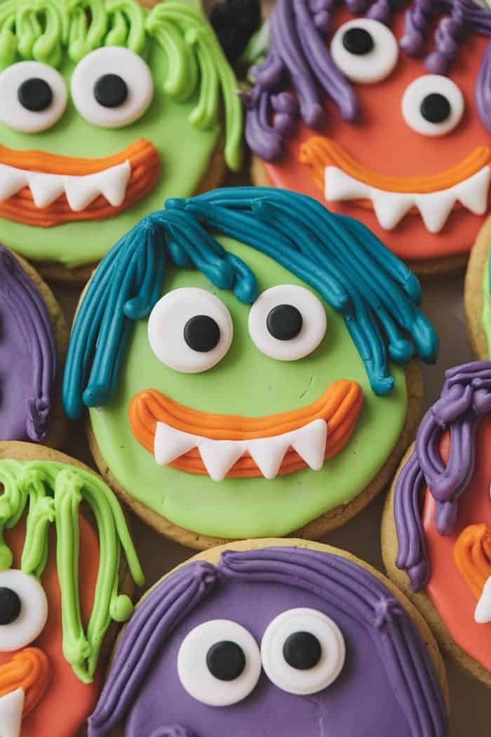 Colorful monster-themed cookies with different faces. Each cookie has bright icing featuring large eyes, jagged teeth, and playful hairstyles in green, blue, orange, and purple. The cookies are arranged closely together.