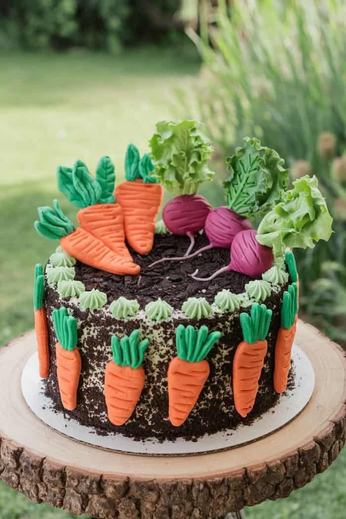 A whimsical cake designed to look like a garden, featuring fondant carrots and beetroots on top and around the sides. The cake is placed on a wooden stand with a grassy garden background.
