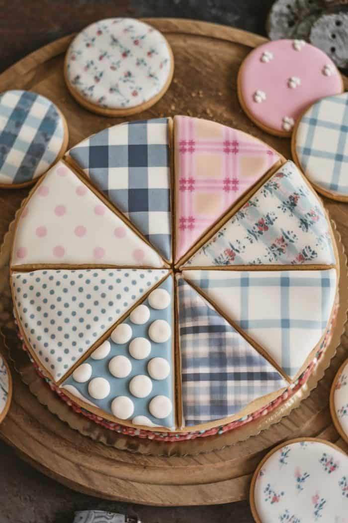A round cake divided into eight slices, each with different plaid and polka dot icing patterns in pastel colors. The cake is surrounded by smaller cookies with similar designs on a wooden board.