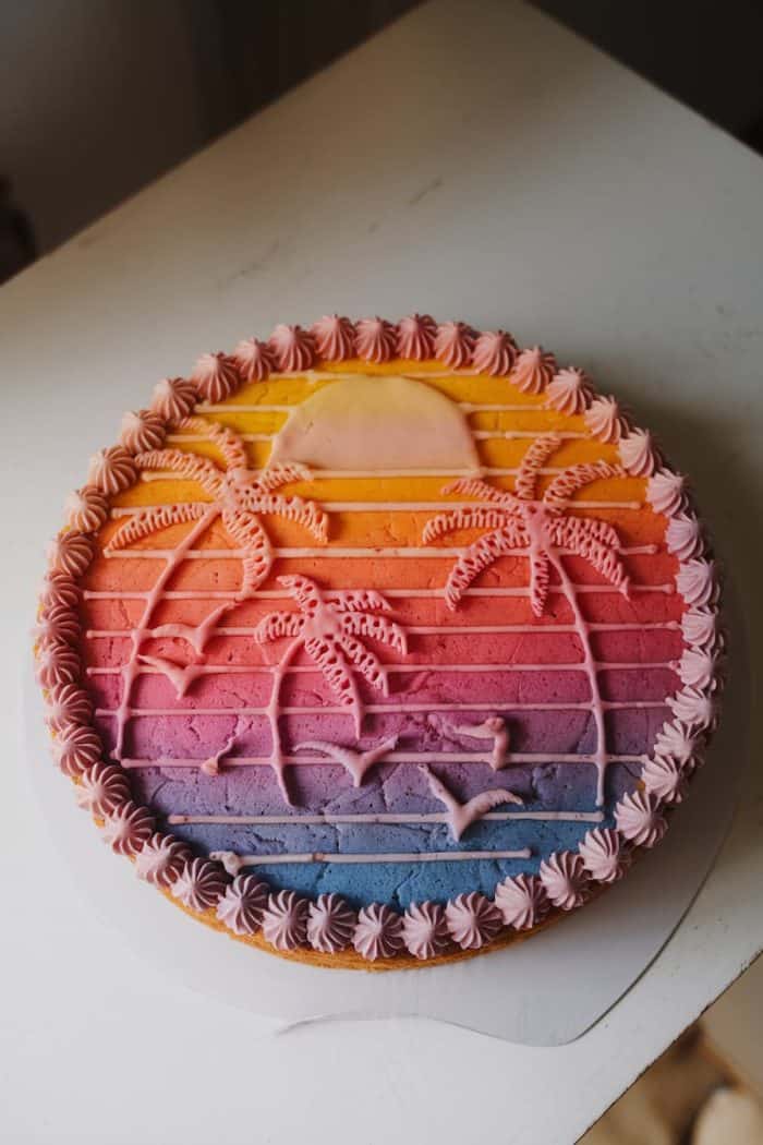 A round cake with a sunset design featuring gradient colors from pink to orange to purple. It is decorated with icing illustrations of palm trees and birds, surrounded by a pink piped border. The cake sits on a white surface.