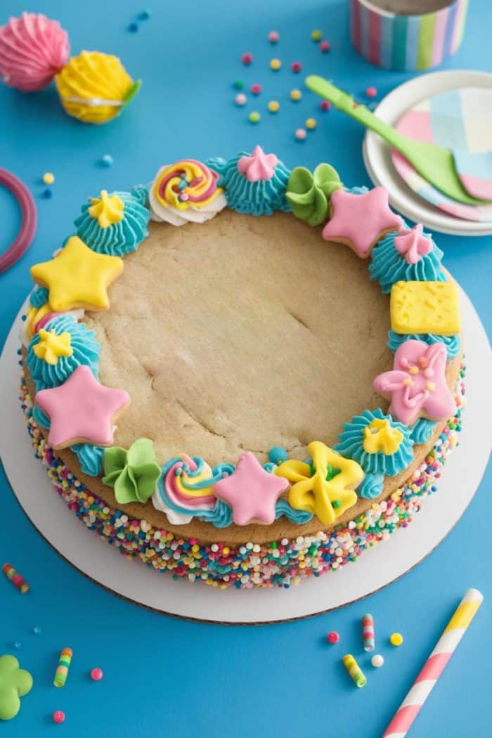 A decorated cake on a blue surface, featuring colorful frosting stars, spirals, and other shapes around the edge. The sides are coated with rainbow sprinkles. Plates and utensils are in the background.