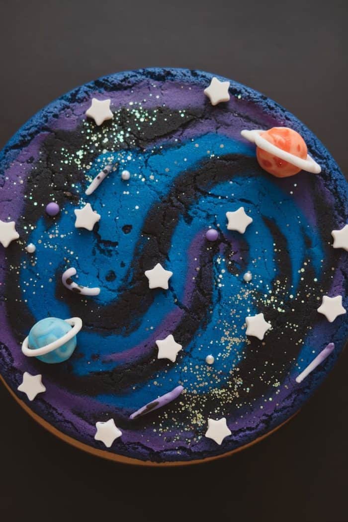 A round cake decorated with a galaxy theme features swirls of blue and black icing. White star-shaped fondants and candy planets are scattered on top, along with small sugar pearls and glitter, creating a celestial appearance against a dark background.