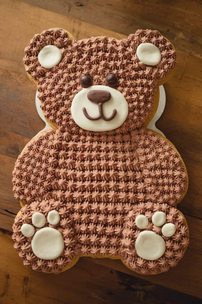 A cake shaped like a teddy bear with chocolate frosting. The bear has white frosting accents on its face, ears, and paws, and chocolate decorations for its eyes, nose, and mouth. It's displayed on a wooden surface.