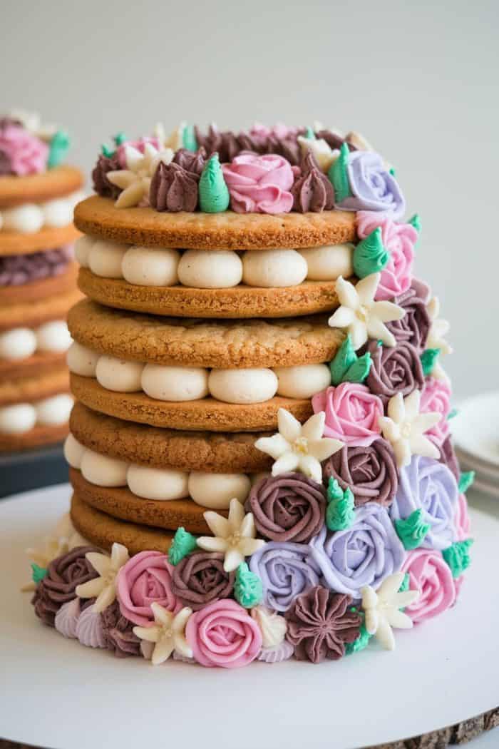 Tall layered cake adorned with alternating cream filling and decorated with colorful flowers in shades of pink, purple, and white. The flowers are artfully arranged on the sides and top, adding elegance to the dessert.
