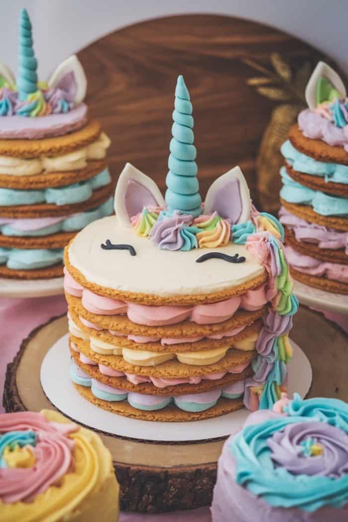 A whimsical unicorn-themed cake with multiple layers, decorated with pastel-colored frosting, includes a spiral horn, ears, and a mane in shades of blue, pink, yellow, and purple. The cake is placed on a wooden platter, surrounded by similar smaller cakes.