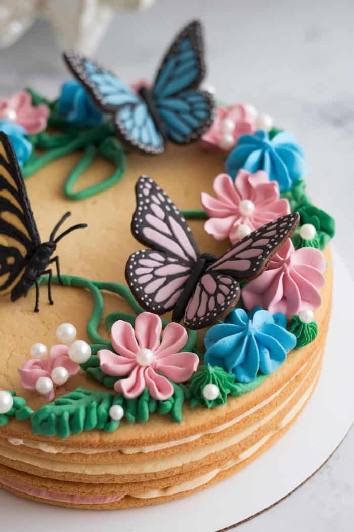 A layered cake decorated with pink and blue frosting flowers, green leaves, and edible pearls. Three edible butterfly toppers in pink, blue, and black sit on top, creating an elegant and colorful design.