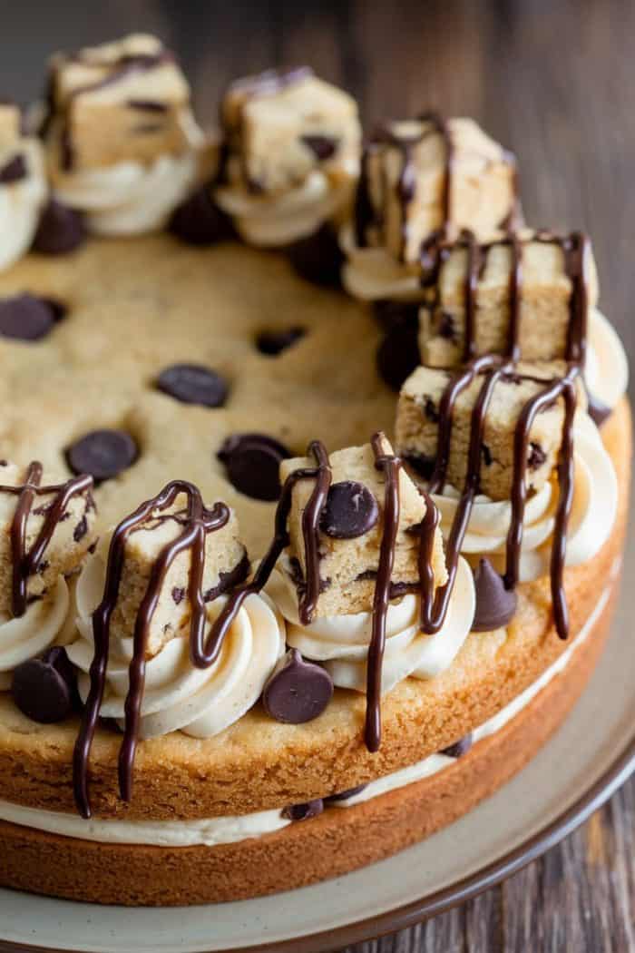 A layered chocolate chip cookie cake topped with swirls of frosting, chocolate chips, cookie pieces, and drizzled chocolate. The cake is displayed on a textured wooden surface.