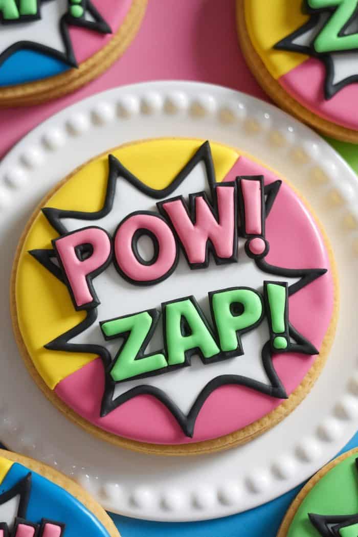 A colorful cookie decorated with comic book-style icing. The words "POW!" and "ZAP!" are in pink and green, respectively, against a yellow and pink background, featuring a white burst outline. Other similar cookies are partially visible.