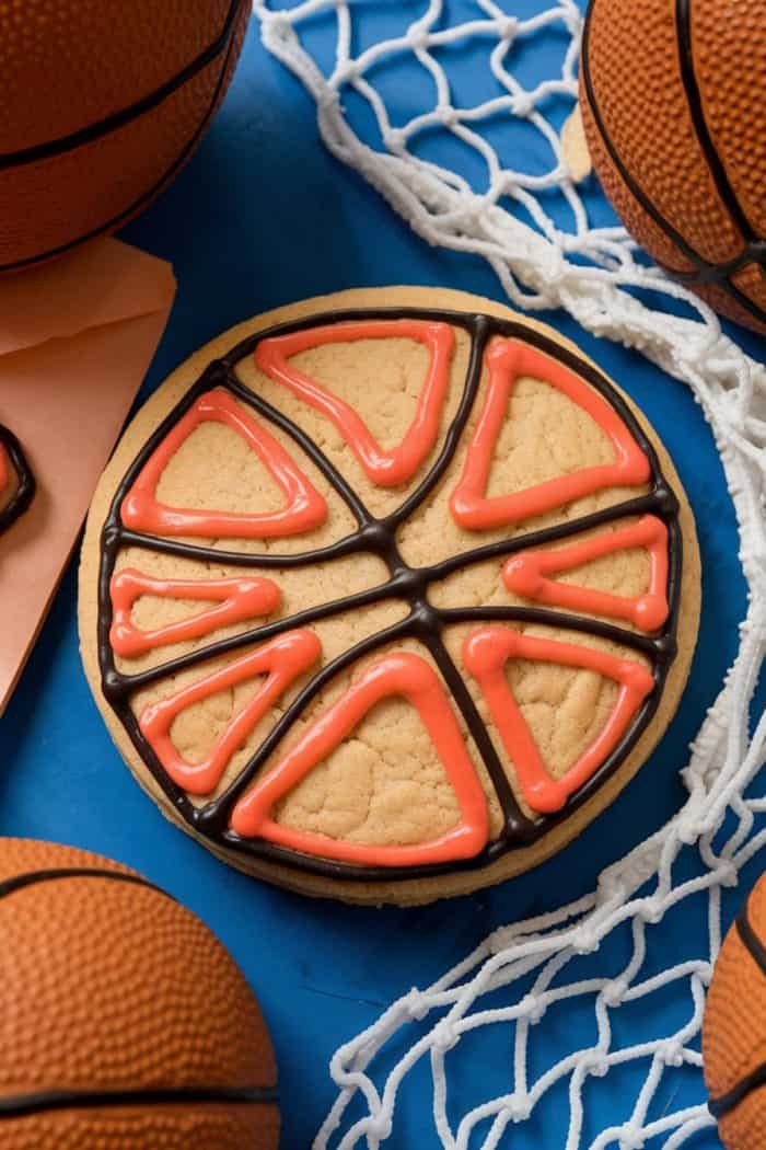 A cookie decorated to resemble a basketball, featuring orange and black icing forming sections similar to a basketball's pattern. It is surrounded by small basketballs and lying on a blue surface with a basketball net nearby.