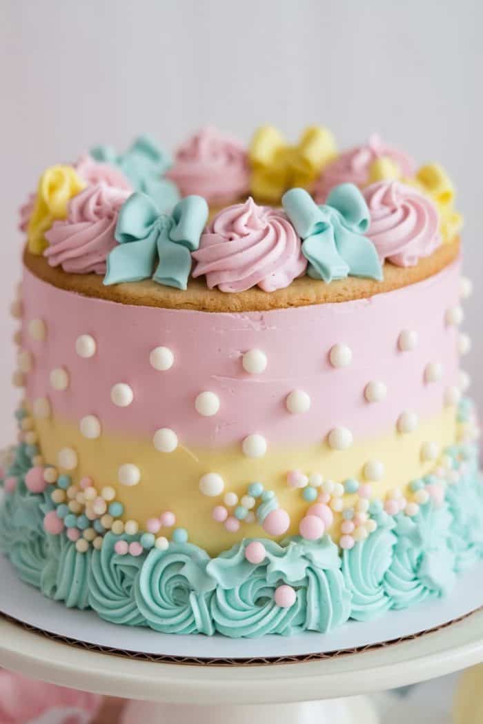A colorful cake with a pink and yellow gradient, decorated with pastel blue and pink frosting swirls, white pearls, and pastel candy balls. The top features pink and blue frosting rosettes with bows.