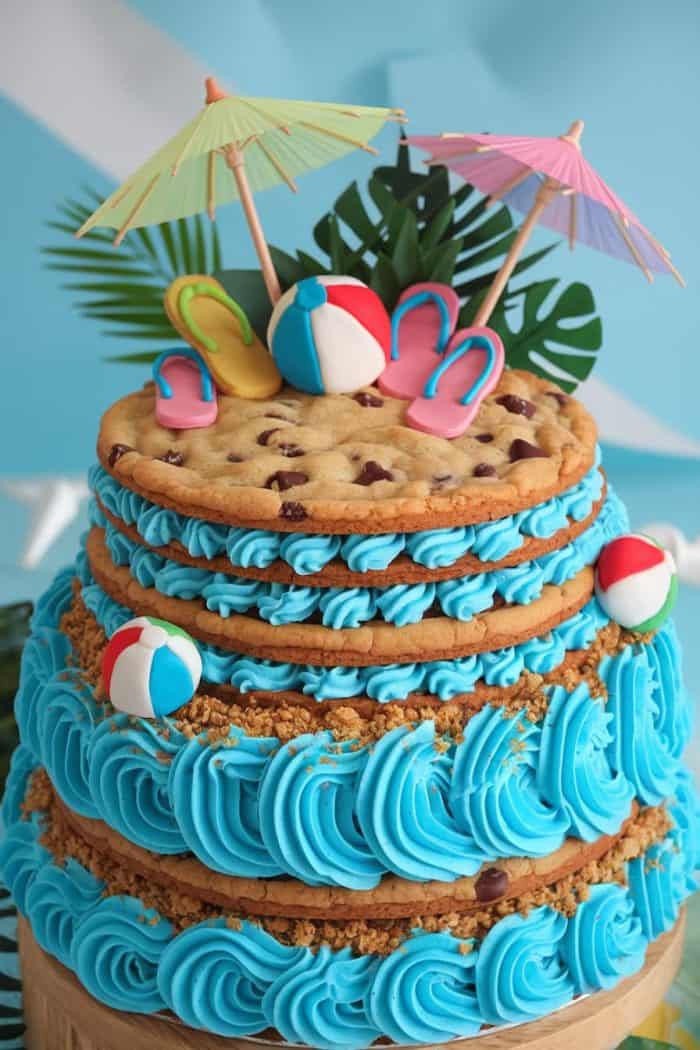 A beach-themed cake with layers of chocolate chip cookies and blue frosting. It is decorated with mini flip-flops, beach balls, and cocktail umbrellas. Tropical leaves are in the background.