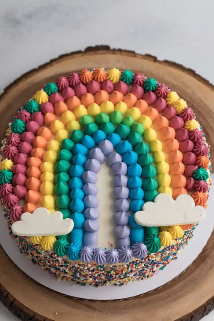 A round cake decorated with rainbow-colored frosting arcs and two white clouds, set on a wooden platter. The sides are covered in multicolored sprinkles.