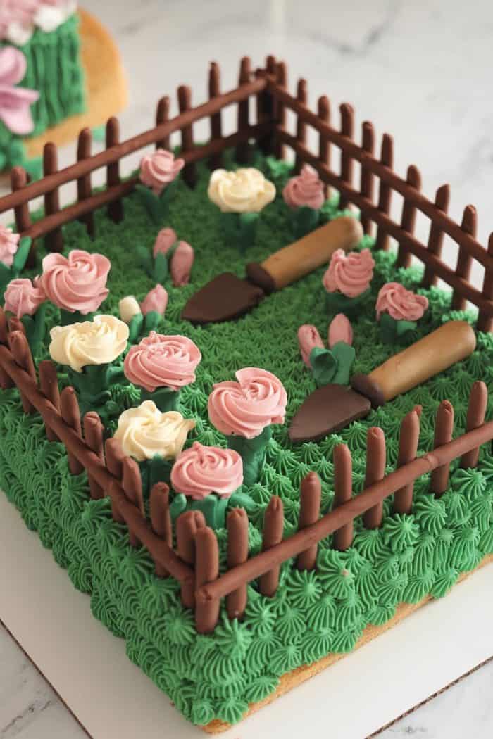 Cake designed as a garden with pink and white icing roses, green icing grass, and chocolate fence. Two small icing trowels lie on the grass, creating a realistic garden scene on a marble background.