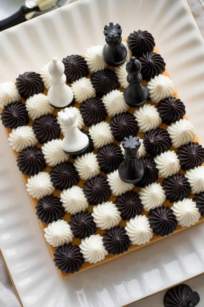 A rectangular cake designed as a chessboard with alternating black and white frosting dollops. Chess pieces, including black and white rooks and a queen, are placed on top. The cake is on a square white plate.