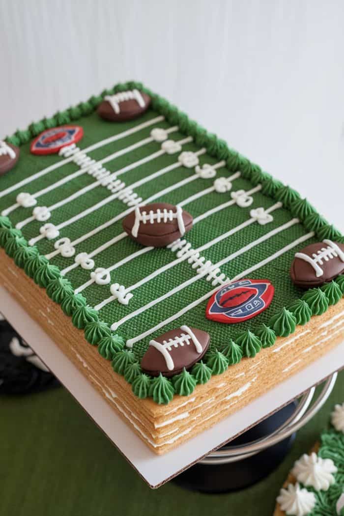 A rectangular cake decorated to resemble a football field, with green frosting as grass and white lines marking the field. Chocolate footballs and red end zone designs are on top, and small white cake flowers border the edges.