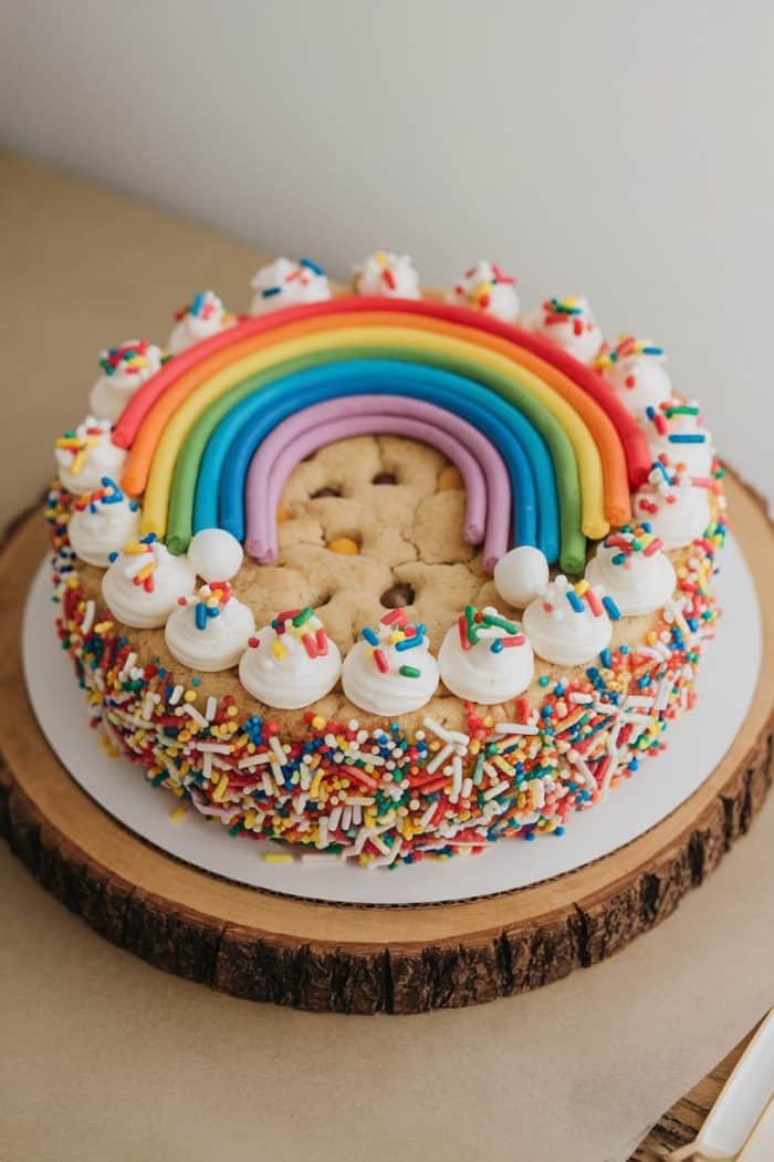A colorful cake decorated with a large fondant rainbow on top, surrounded by whipped cream dollops, each topped with multicolored sprinkles. The sides of the cake are covered in sprinkles, and it rests on a wooden slab.