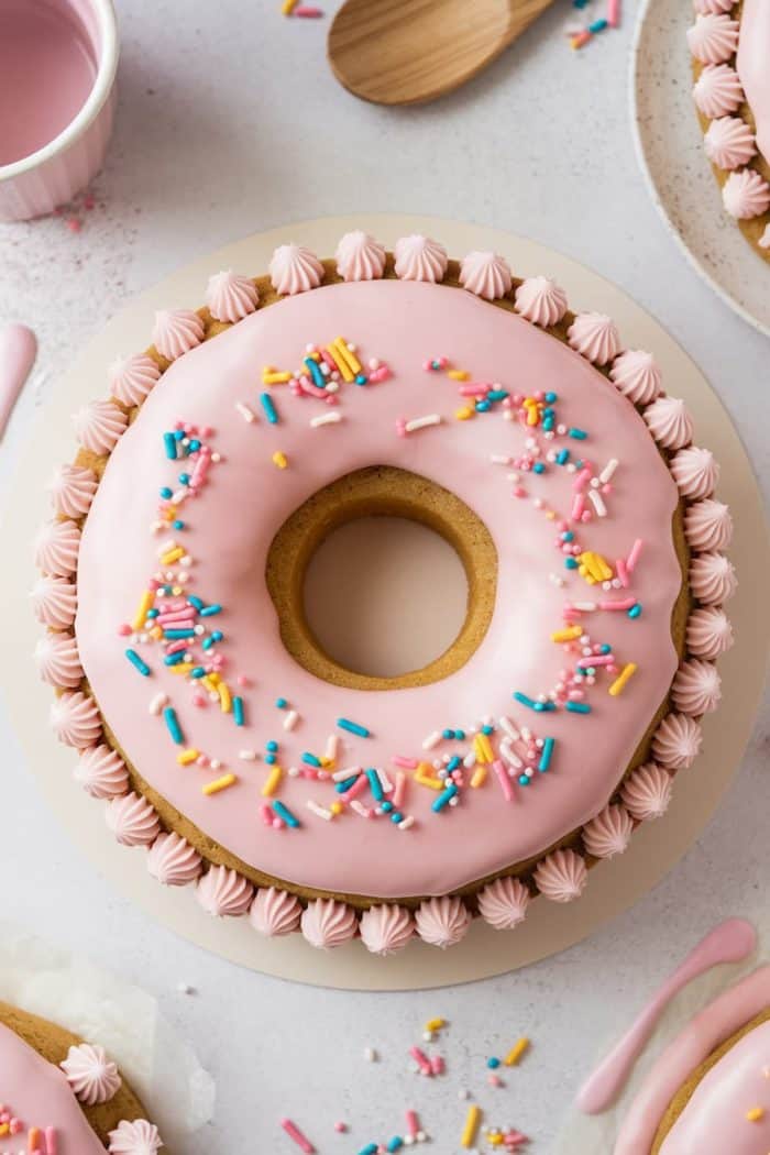 A round cake with pink frosting and colorful sprinkles, topped with small pink icing rosettes along the edge. A wooden spoon is nearby on a light surface.