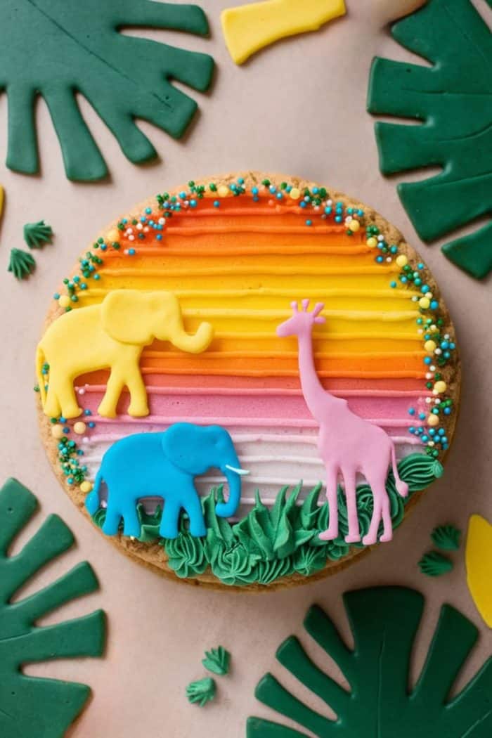 Round cookie with a rainbow-striped frosting background, topped with colorful decorations of an elephant, giraffe, and leaves. Surrounded by green fondant leaves and small decorative elements.