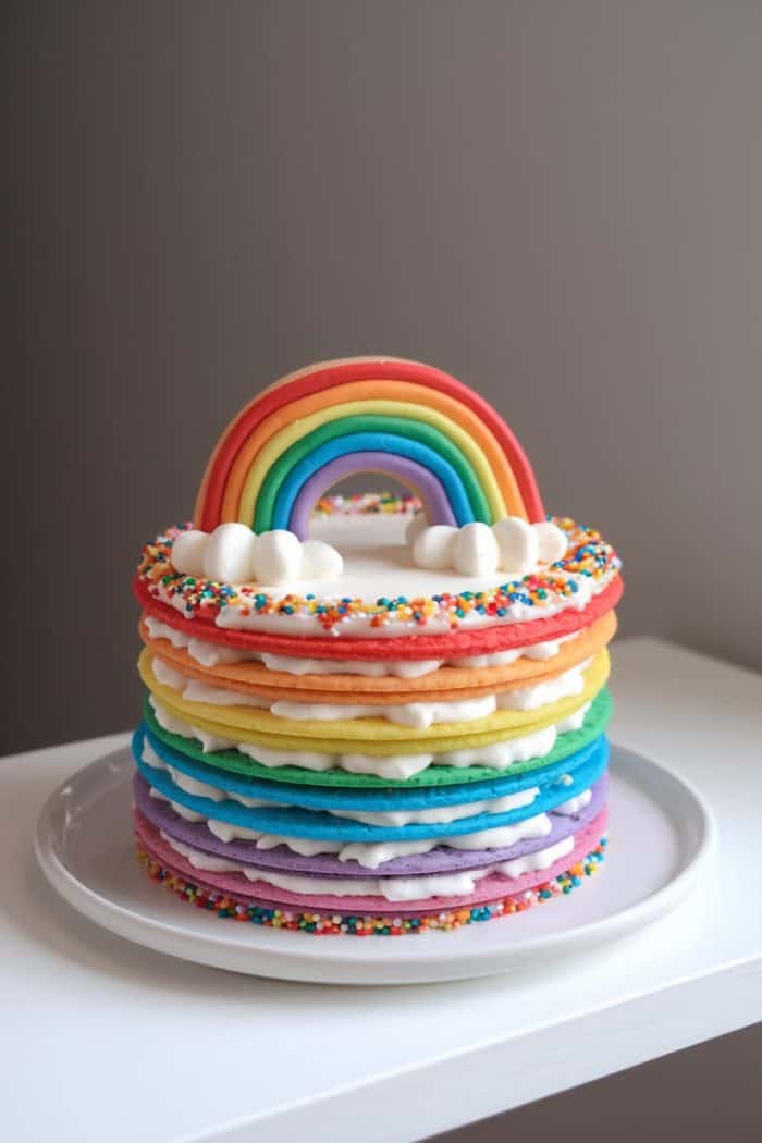 A colorful cake with multiple rainbow-colored layers, topped with white frosting and sprinkles. A fondant rainbow arches over the top, resting on small white cloud-shaped decorations. The cake is set on a white plate.