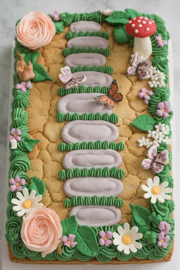 A rectangular cake decorated to resemble a garden path with stone-like frosting, green grass, and various flowers. It features butterflies, a mushroom, roses, daisies, and small white blossoms, creating a whimsical garden scene.