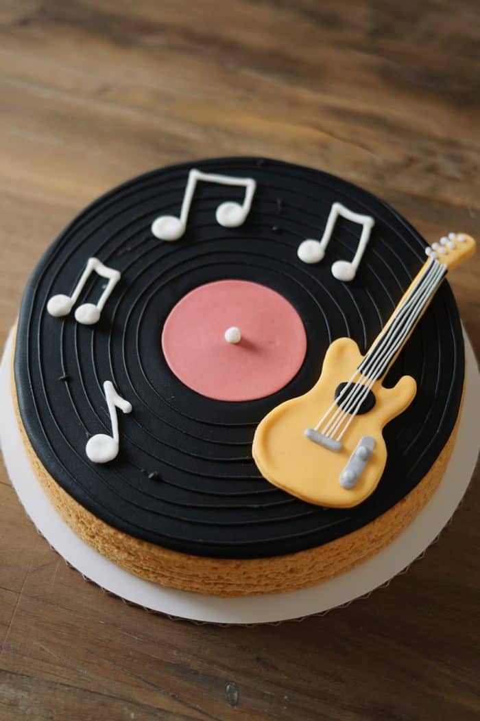 A cake decorated to resemble a vinyl record with music notes and a yellow guitar on top, sitting on a wooden table. The cake has a black fondant surface with a pink center.
