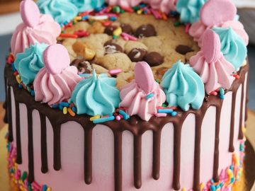A colorful cake with pink frosting and chocolate drips, topped with blue and pink swirls, pink candies, and a large cookie. Rainbow sprinkles decorate the base. The cake is on a brown board.