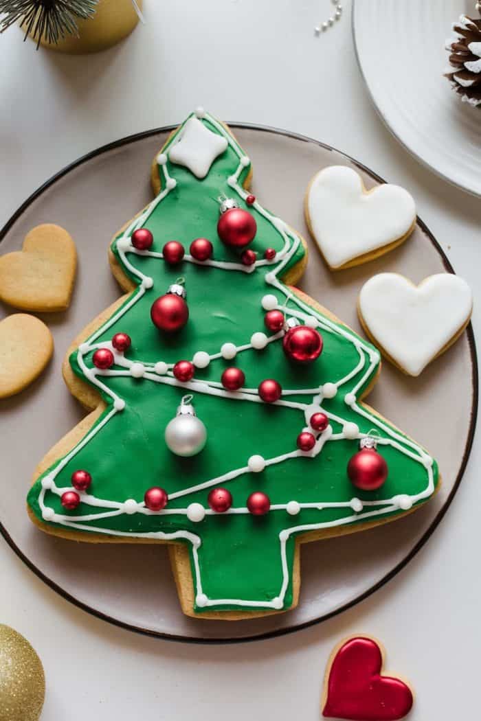A large Christmas tree-shaped cookie decorated with green icing, white frosting garlands, and red and white candy ornaments on a plate. Surrounding it are small heart-shaped cookies, some plain and others with white icing.