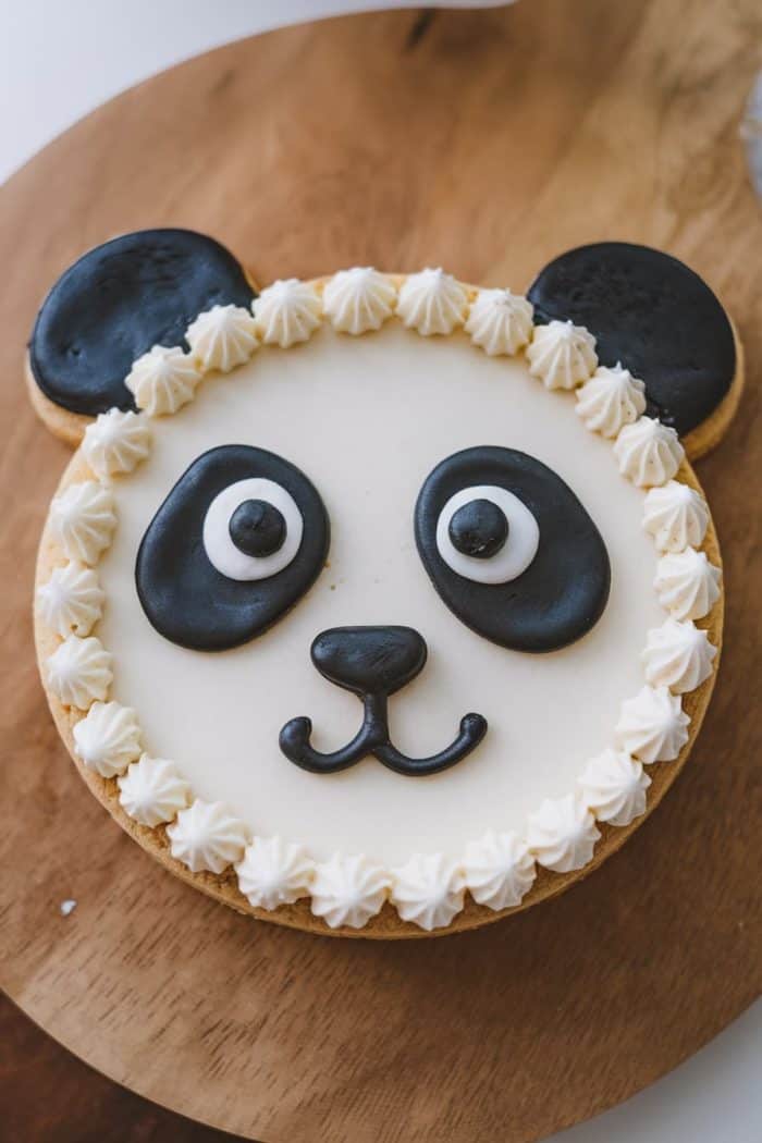 A panda-themed cake on a wooden surface, decorated with white frosting and black fondant resembling panda eyes, ears, and nose, with piped cream around the edge.