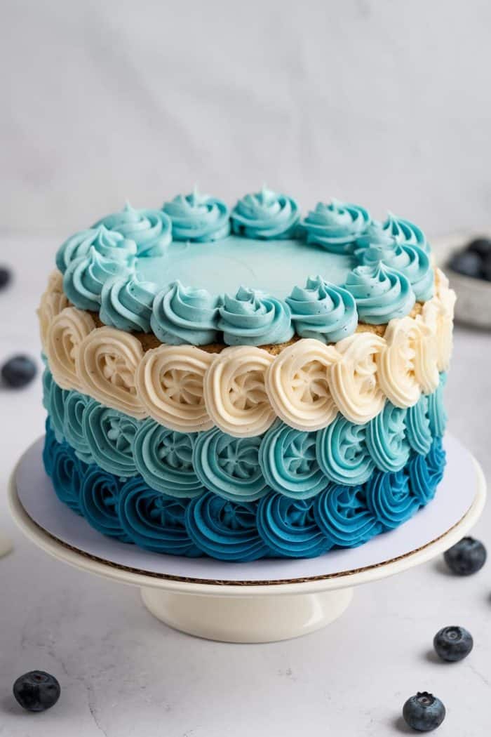A three-tiered cake with ombre blue and cream frosting is decorated with piped swirls. The bottom layer is deep blue, the middle is cream-colored, and the top is light blue. Blueberries are scattered around the white cake stand on a marble surface.