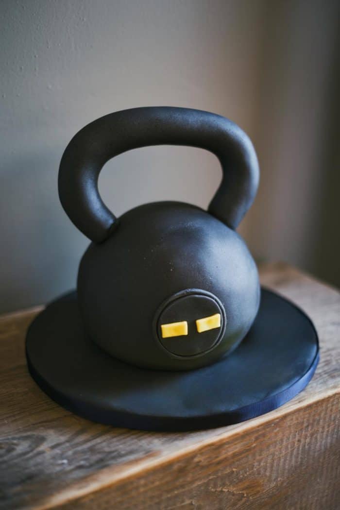 A cake shaped like a black kettlebell sits on a wooden surface. It has a circular base and small yellow details on the front, resembling a handle weight. The background is a plain wall.
