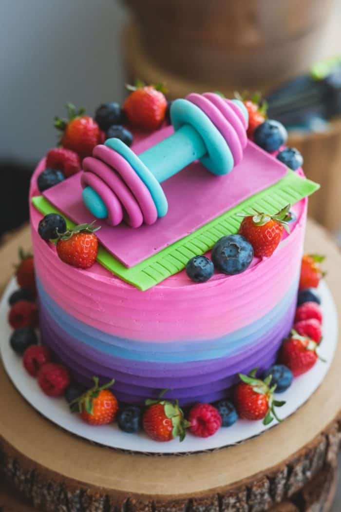 A colorful cake with pink and purple gradient icing, topped with a small teal and pink dumbbell decoration. The cake is adorned with strawberries, blueberries, and raspberries around the base.