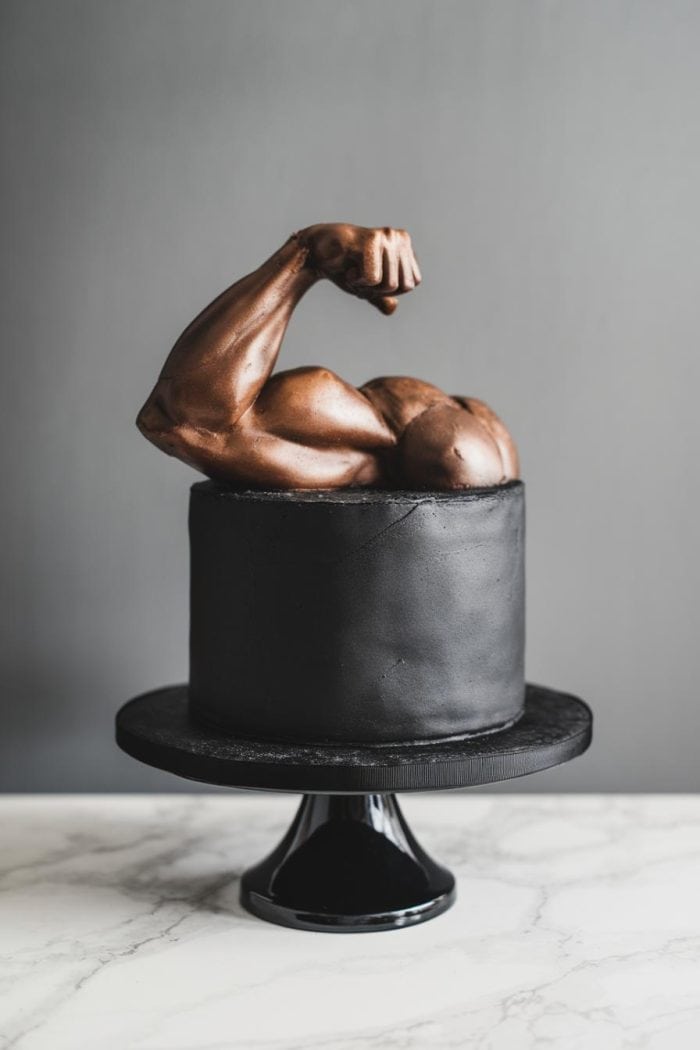 A black cake with a sculpted arm flexing a bicep on top, resembling a muscular bronze figure, sitting on a black cake stand against a gray background.