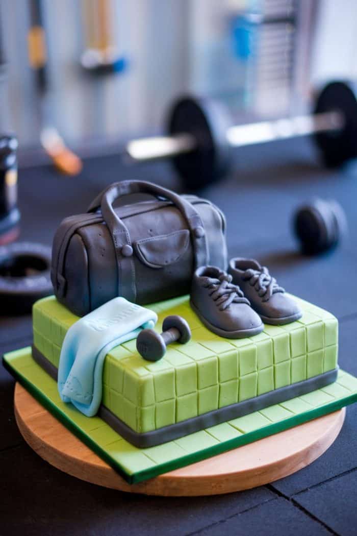 A fitness-themed cake with a green base features a fondant gym bag, sneakers, a dumbbell, and a towel. In the blurred background, gym equipment and weights are visible.