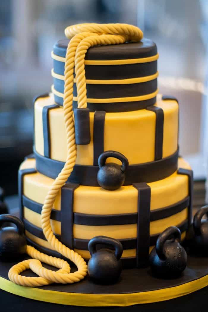 A three-tiered cake decorated in yellow and black fondant, designed to look like weights. A fondant rope drapes over the top tier, and small kettlebell decorations surround the cake on the base.