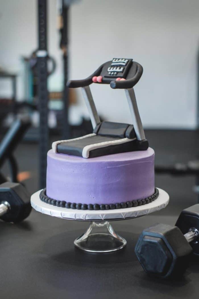 A purple cake with a treadmill decoration on top is displayed on a glass stand. Nearby, dumbbells are visible, suggesting a fitness or workout theme.