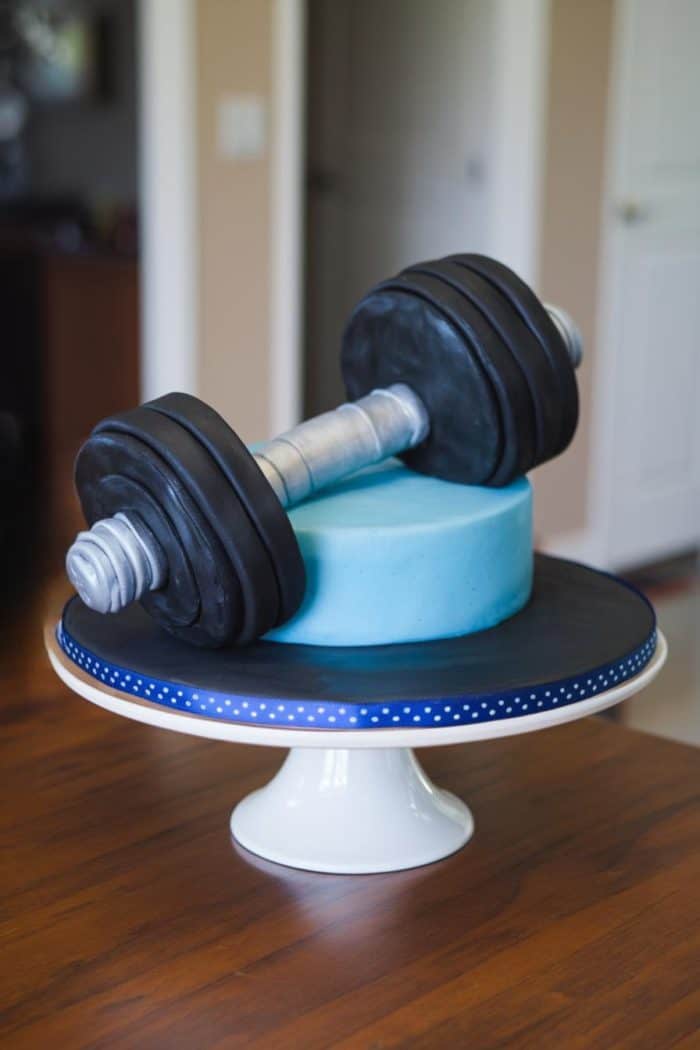 A round cake decorated with blue icing sits on a white cake stand with a blue polka dot ribbon. On top of the cake is a realistic-looking dumbbell made of black and silver fondant.