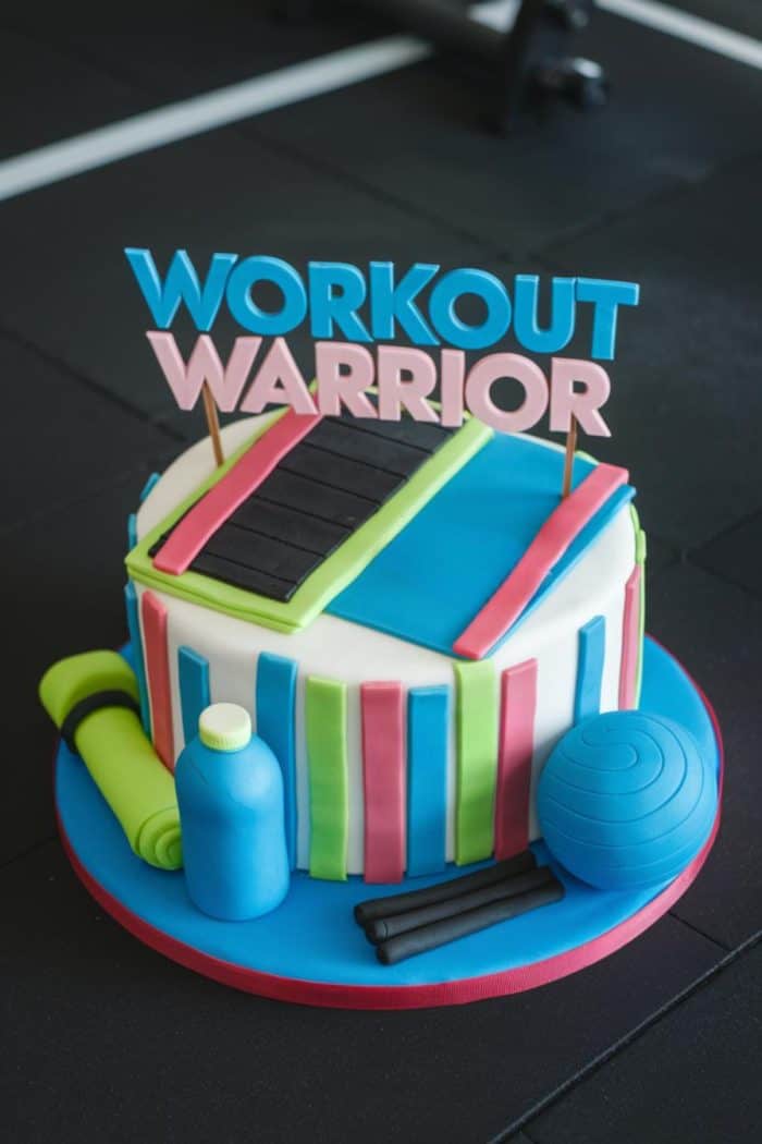 A round cake decorated with gym-themed fondant elements like a yoga mat, water bottle, exercise ball, and dumbbells. The cake is topped with the words "Workout Warrior" in bold blue and pink letters.