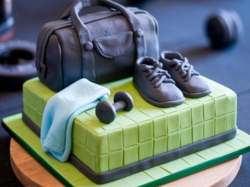 A rectangular cake decorated with a black fondant gym bag, a pair of sneakers, a small dumbbell, and a light blue towel draped over the side. The cake is green with a grid pattern, and sits on a round wooden board.