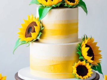 A two-tiered cake decorated with yellow and white stripes and adorned with realistic sunflower decorations. The cake sits on a wooden stand, surrounded by additional sunflower accents on a marble surface. A fork and knife are placed nearby.