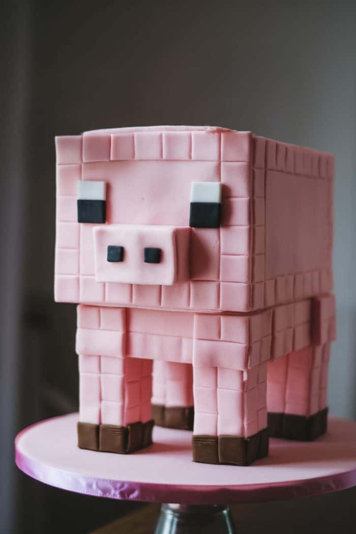 A cake designed to look like a pixelated pink pig, reminiscent of a video game style. It features blocky shapes with black and white square eyes and a pink square nose, standing on a pedestal with a light pink base.