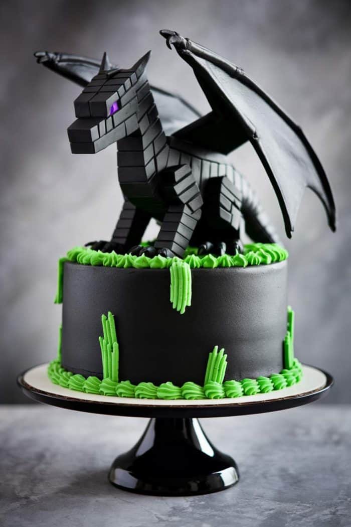 A black cake topped with a detailed dragon figure. The cake has smooth black icing and is decorated with neon green icing accents resembling spikes. The dragon has purple eyes and spread wings, sitting on a round pedestal.
