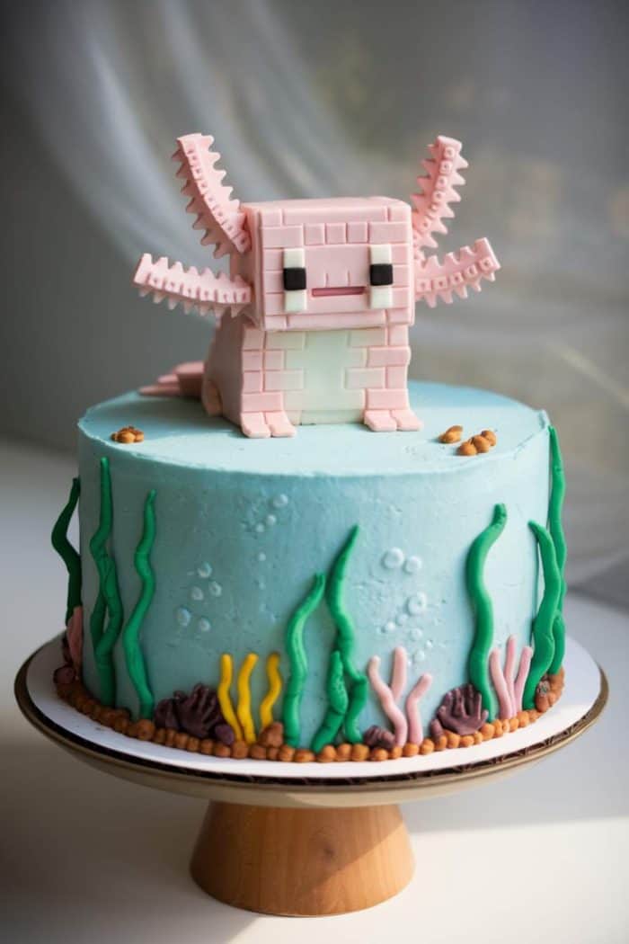 A cake decorated with light blue frosting and an undersea theme. Green and yellow fondant sea plants line the base. On top is a pink, blocky axolotl figure with squared details, resembling a Minecraft style.
