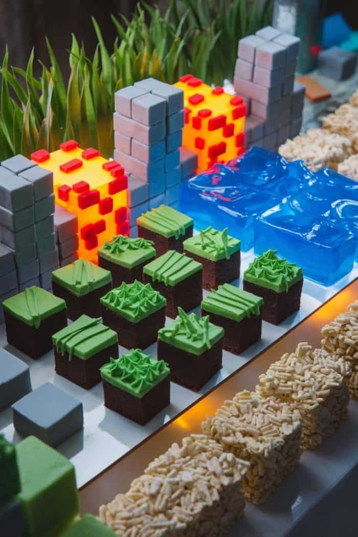 An assortment of Minecraft-themed desserts, including grass block cakes, blue jelly water, glowing lava cube confections, and rice cereal blocks, are arranged on a white table. Tall grass decor enhances the theme.