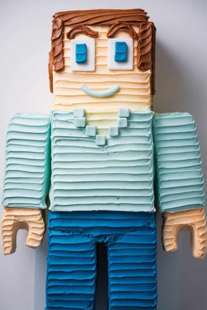 A cake designed to look like a pixelated character with brown frosting hair, a blue and white square-eye face, a teal shirt with a detailed collar, and blue pants. The frosting is applied in a textured, striped pattern.