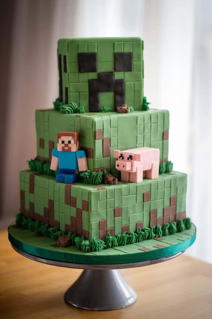 A three-tiered Minecraft-themed cake featuring block designs, a character figure, a pig, and a creeper on top. The cake is decorated in shades of green and brown with small accents resembling grass and rocks, displayed on a metal stand.